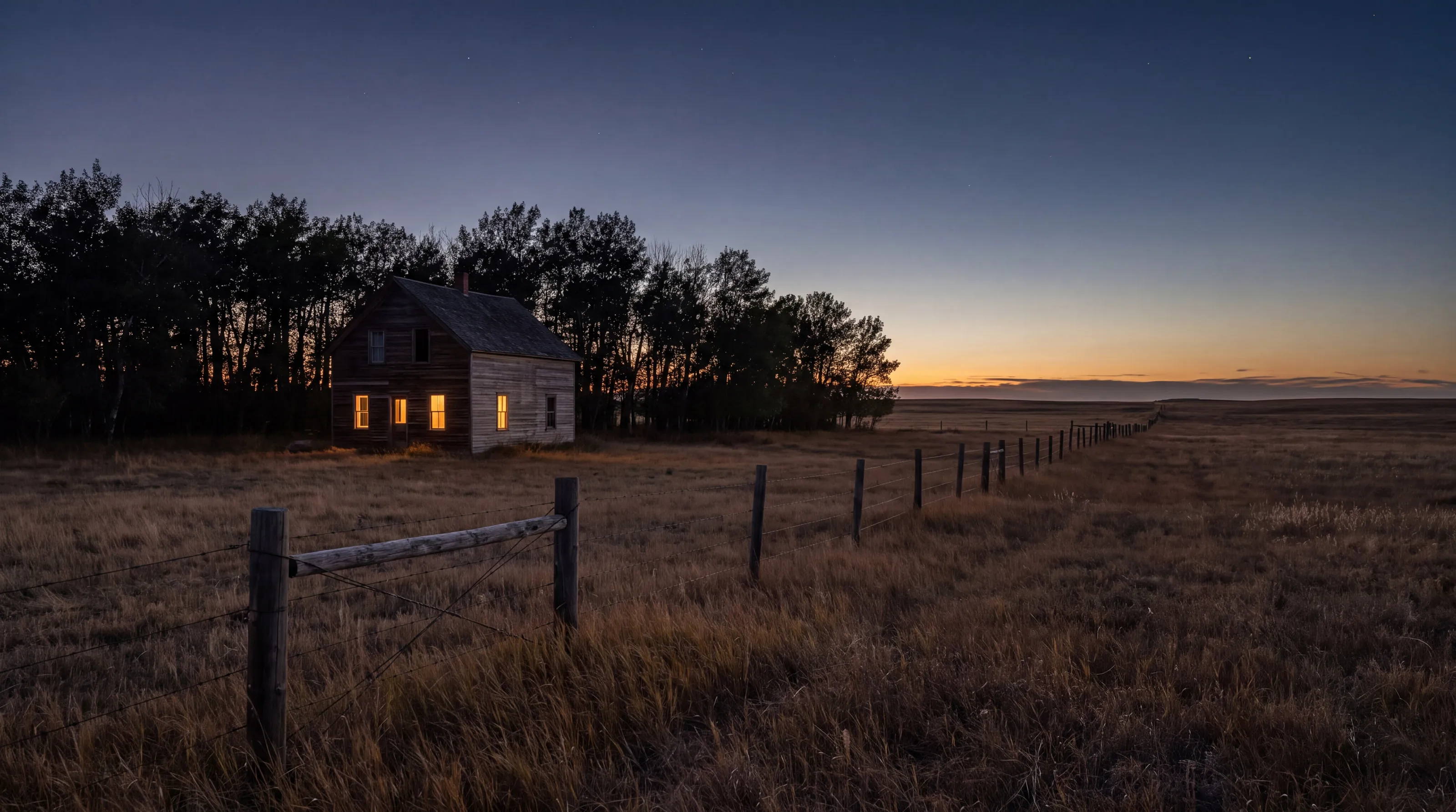 A lone farmhouse with lit windows on the open prairie at dusk